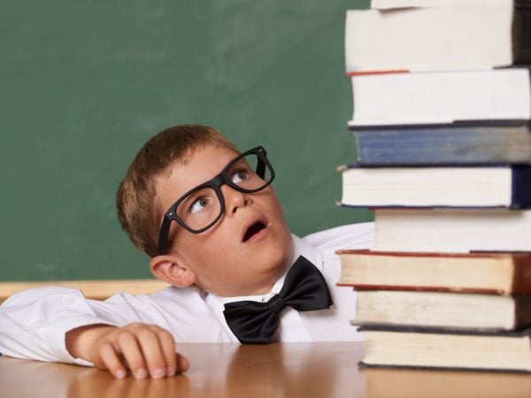 A young boy wearing glasses and a bow-tie looking wearily at a large pile of books